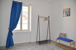 Ein Zimmer mit Fenster, einem blauen Vorhang, einem schwarzen Metallgestell mit Kleidung, einer Holzbank mit blauem Kissen und einem an der Wand hängenden Bild.