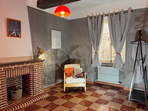 Ein Zimmer mit Kamin aus Backsteinen, einem Sessel, einem Fenster mit gestreiften Vorhängen und einer Hängelampe.
