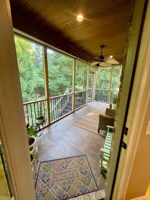 Eine überdachte Veranda mit Blick auf einen Wald, eine Fußmatte und ein Deckenventilator.