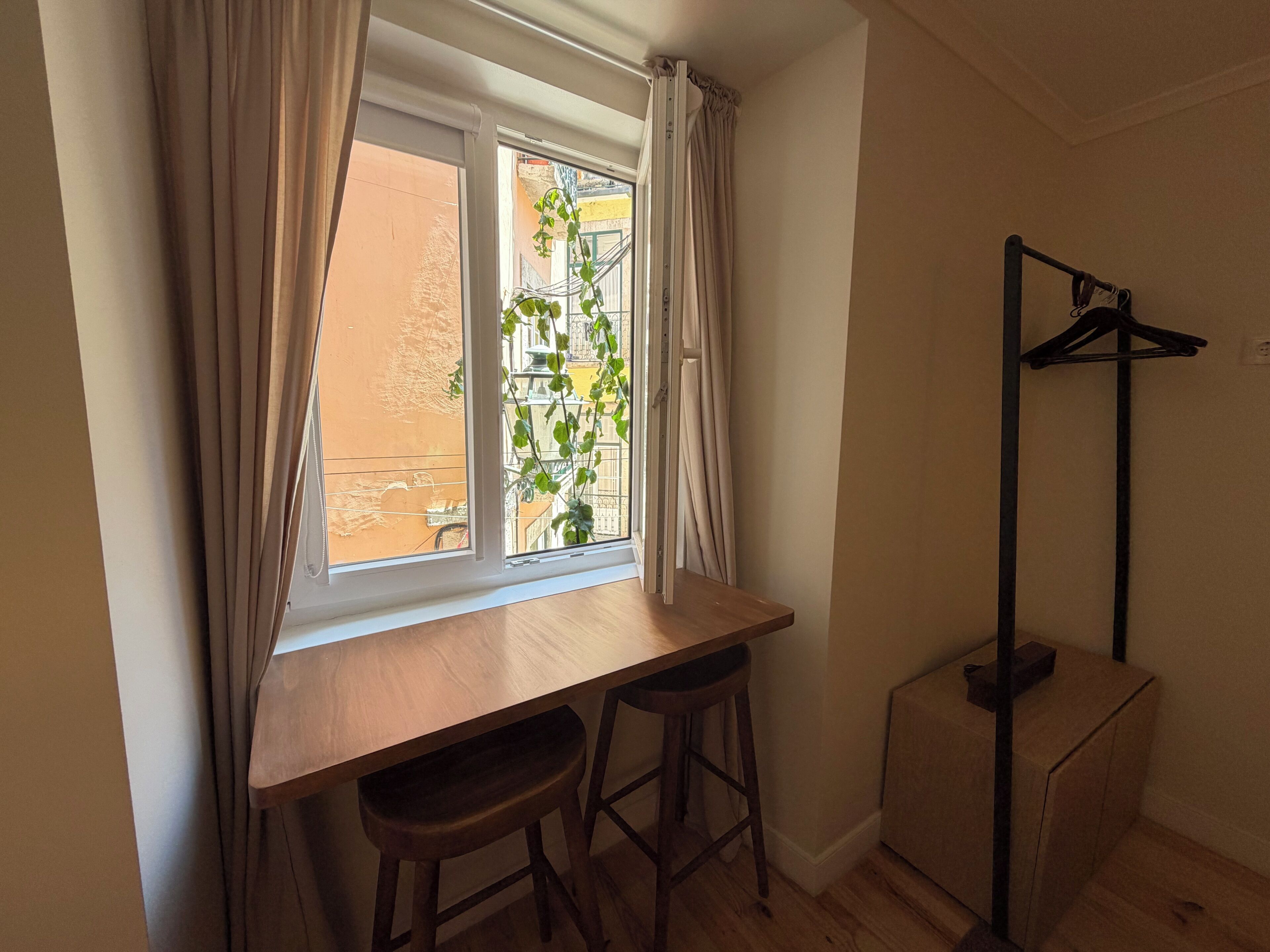 Une pièce avec une fenêtre, un bureau en bois, deux tabourets, un porte-manteau et un petit meuble.