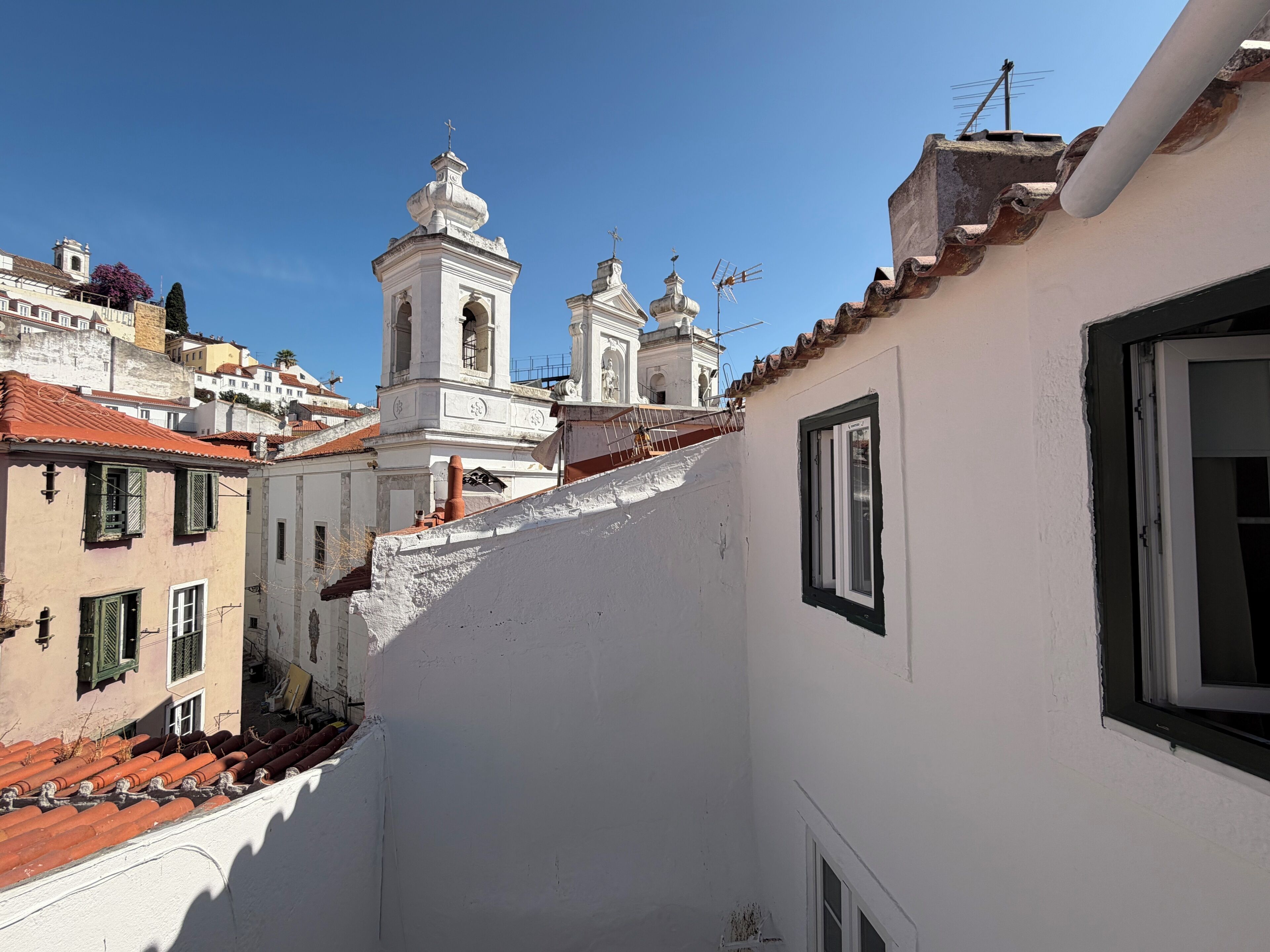 A narrow street with buildings, including a white building with a window and a bell tower in the background.