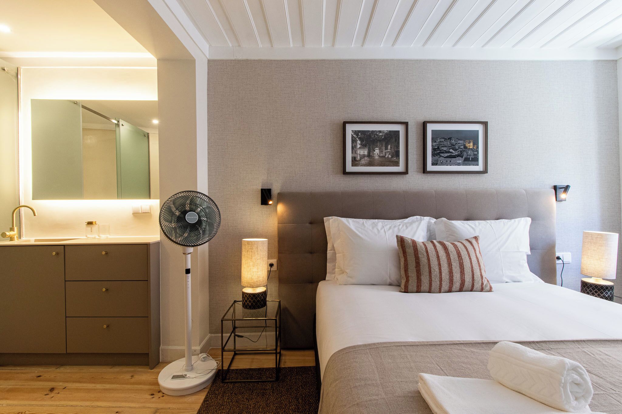 A hotel room with a bed, bedside lamps, a standing fan, a mirror, and a view of the city.