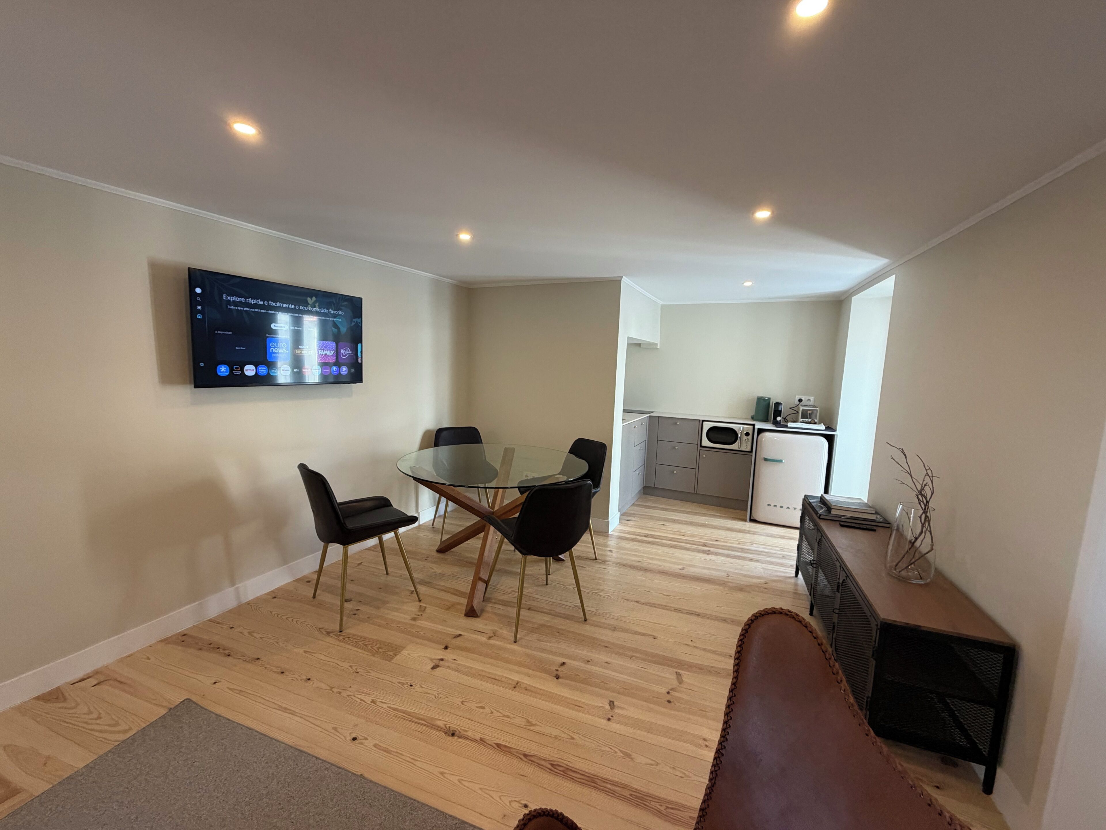 A modern living space with a glass dining table, black chairs, a flat-screen TV mounted on the wall, a kitchenette, and a small side table with a vase.