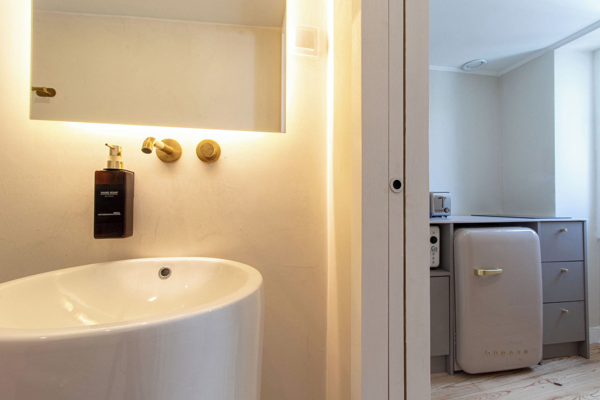 Un bagno moderno con un lavandino bianco grande, un dispenser per sapone e uno specchio. Si vede un piccolo armadietto bianco con un tostapane e un frigorifero.