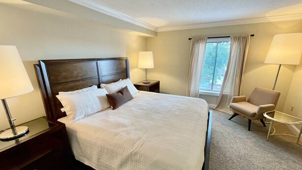 A hotel room with a large bed, two bedside tables with lamps, a chair, and a window with curtains. - Stylish 2 Bedroom Apartment Downtown High Point (High Point)