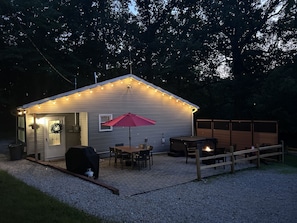 A small, single-story building with a porch, a table, chairs, a red umbrella, and a fire pit.