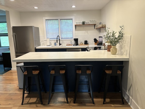A modern kitchen with a large island, bar stools, a refrigerator, and a window with blinds.