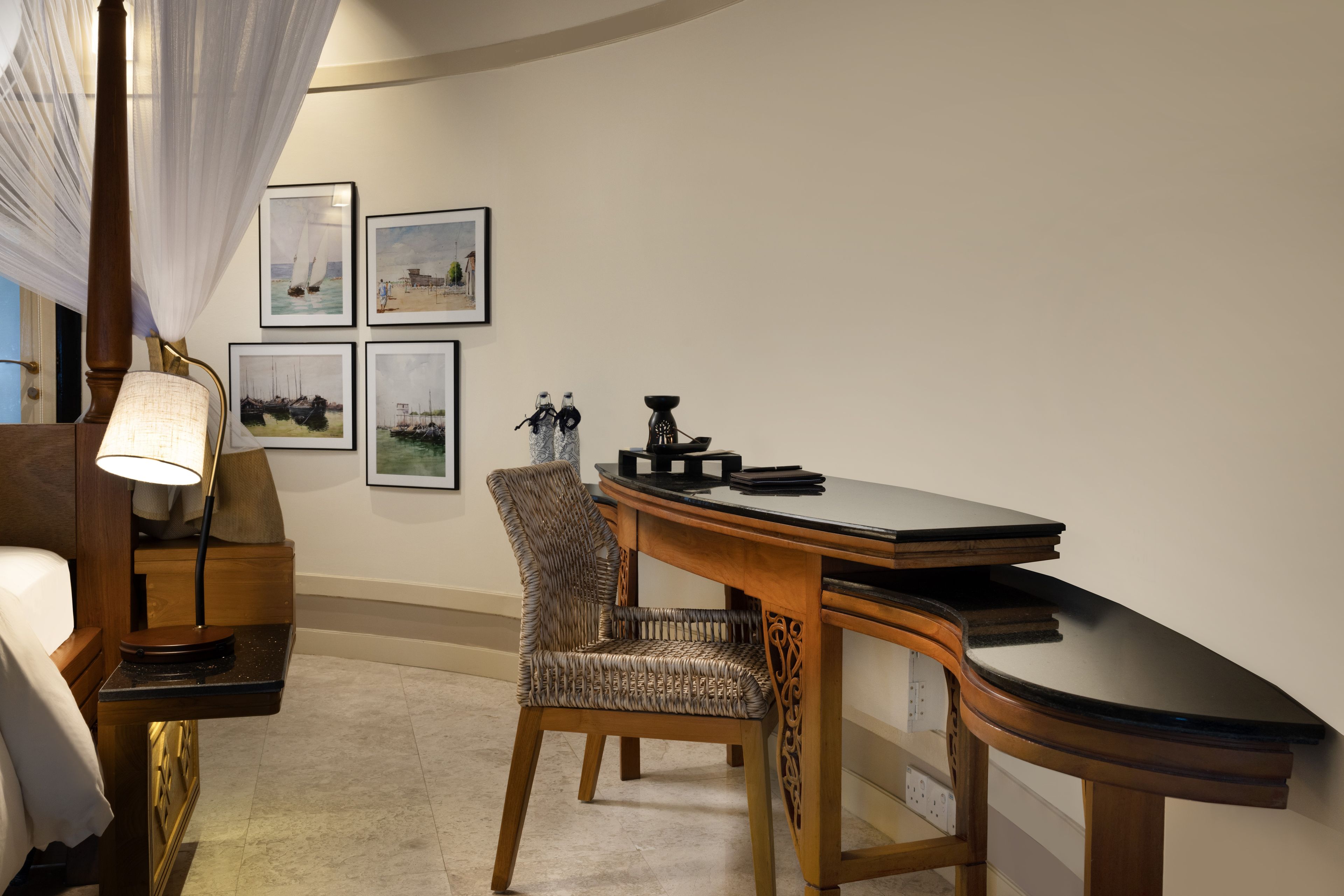 A hotel room with a curved desk, a chair, a lamp, and framed pictures on the wall.