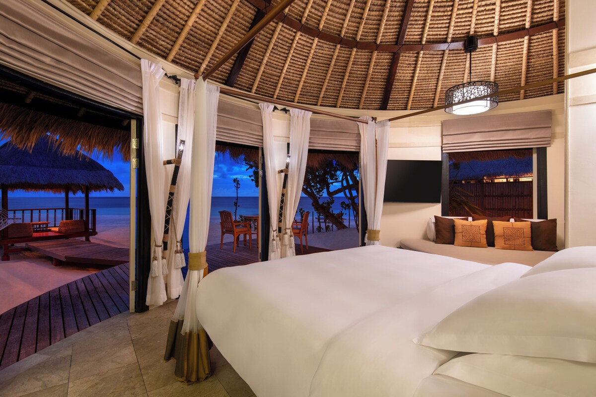 A bedroom with a large bed, a view of the beach, and a thatched roof structure.