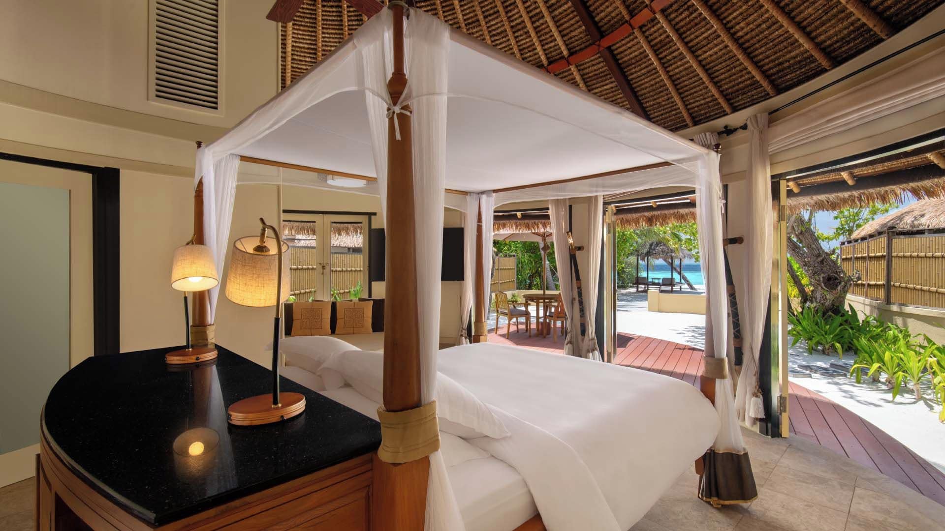 A four-poster canopy bed with a black countertop and a lamp, a television, and a view of the beach and palm trees.