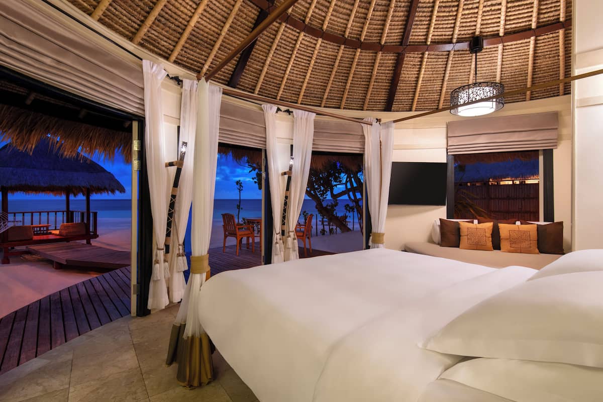 A bedroom with a large bed, a view of the beach, and a thatched roof structure.
