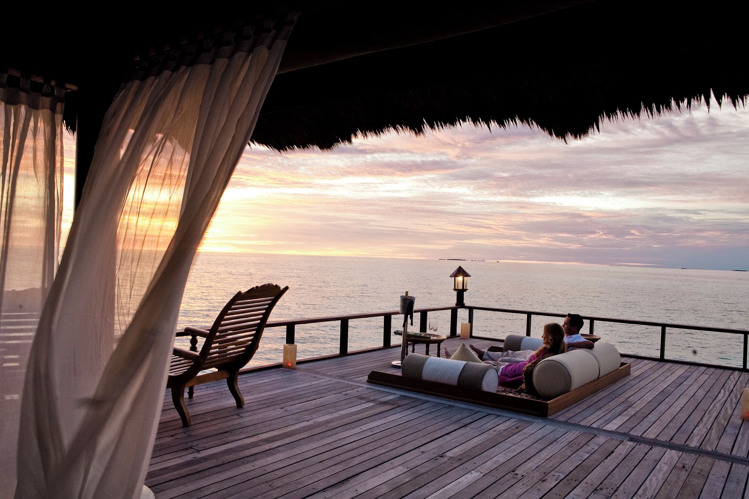 A wooden deck with a couple relaxing, a table with a bottle and glasses, and a view of the sea at sunset.