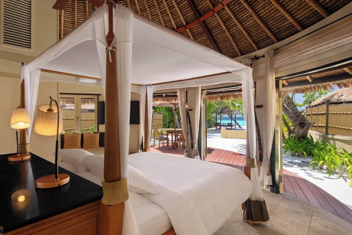 A four-poster canopy bed with a black countertop and a lamp, a television, and a view of the beach and palm trees.