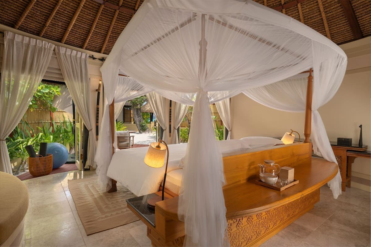 A room with a bed under a canopy, a bedside table with a lamp, and a view of the outdoors.