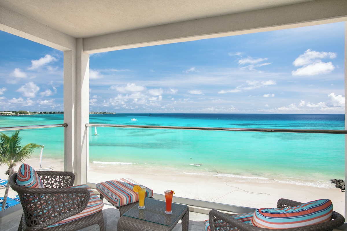 A balcony with wicker furniture, a glass table, and two drinks. The view includes a beach and turquoise waters.
