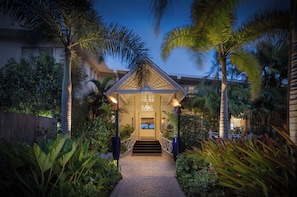 Une entrée bien éclairée, avec un escalier couvert menant à un bâtiment, le tout entouré d’une végétation tropicale luxuriante.