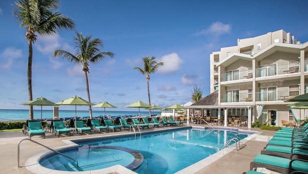 A pool area with lounge chairs and umbrellas, a building with balconies, and palm trees. - Sea Breeze Beach House All Inclusive by Ocean Htls (CHRIST CHURCH)