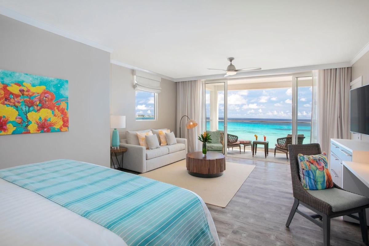 A hotel room with a bed, a sofa, a dining area, and a balcony with a view of the ocean.