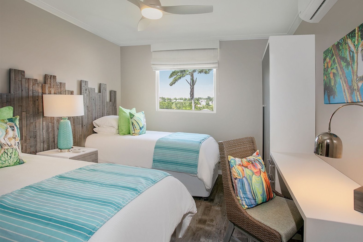 A hotel room with two beds, a wicker chair, a desk, a lamp, and a painting on the wall.