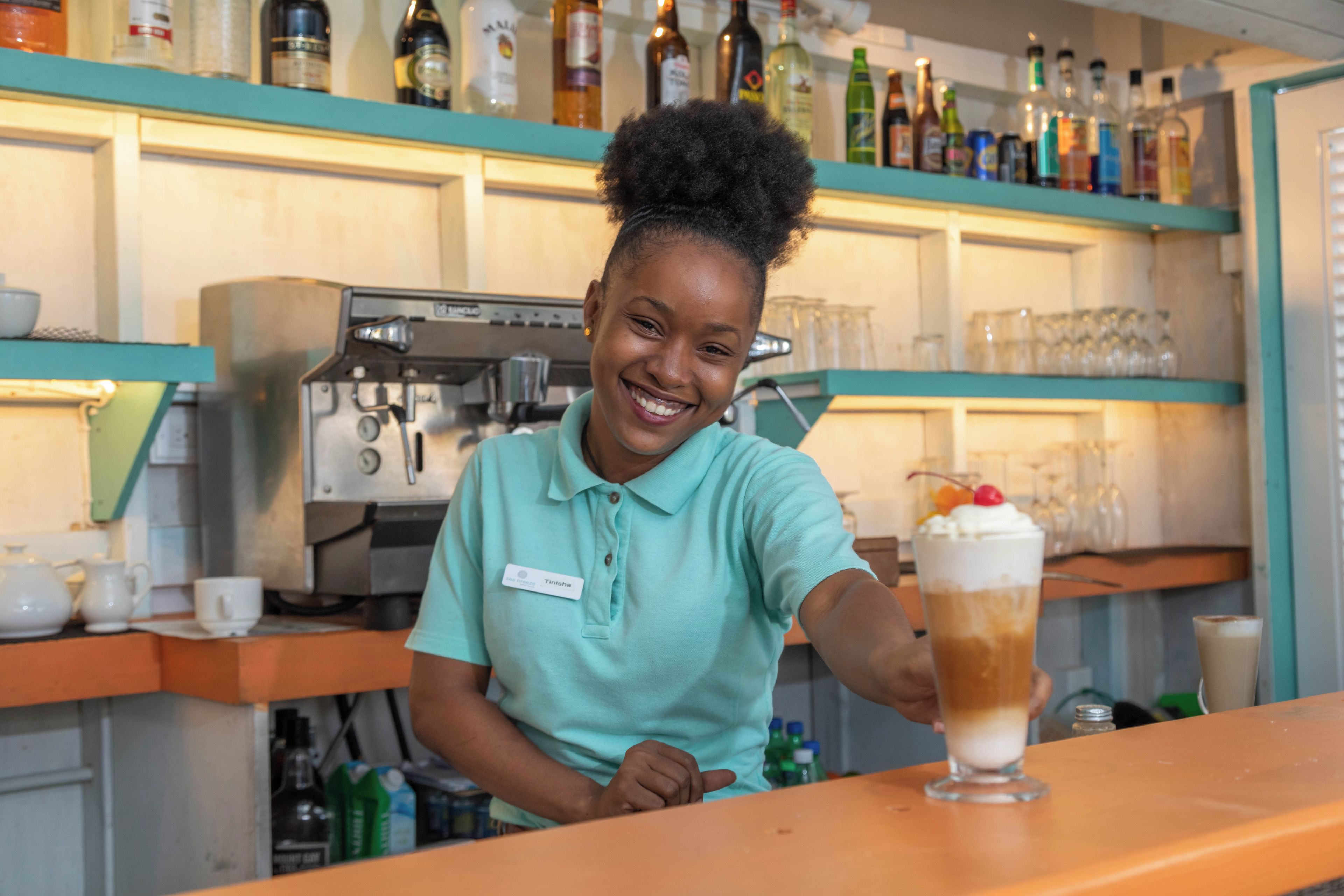A person in a teal shirt behind a bar counter with a beverage in front of them.