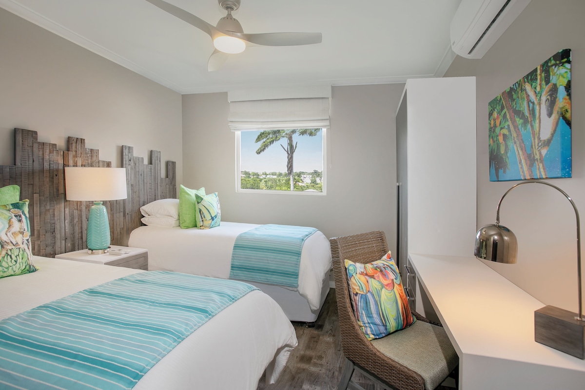 A hotel room with two beds, a window with a view, a ceiling fan, and a painting.