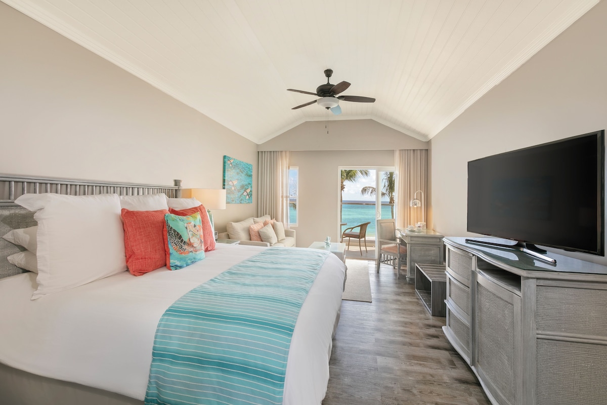A hotel room with a large bed, a TV, a ceiling fan, and a view of the ocean.