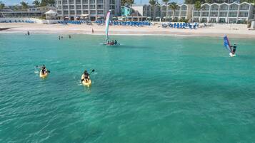 A beach with clear turquoise water, kayaks, a windsurf board, and a hotel with multiple balconies in the background.