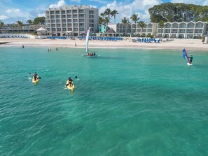 A beach with clear turquoise water, kayaks, a windsurf board, and a hotel with multiple balconies in the background.