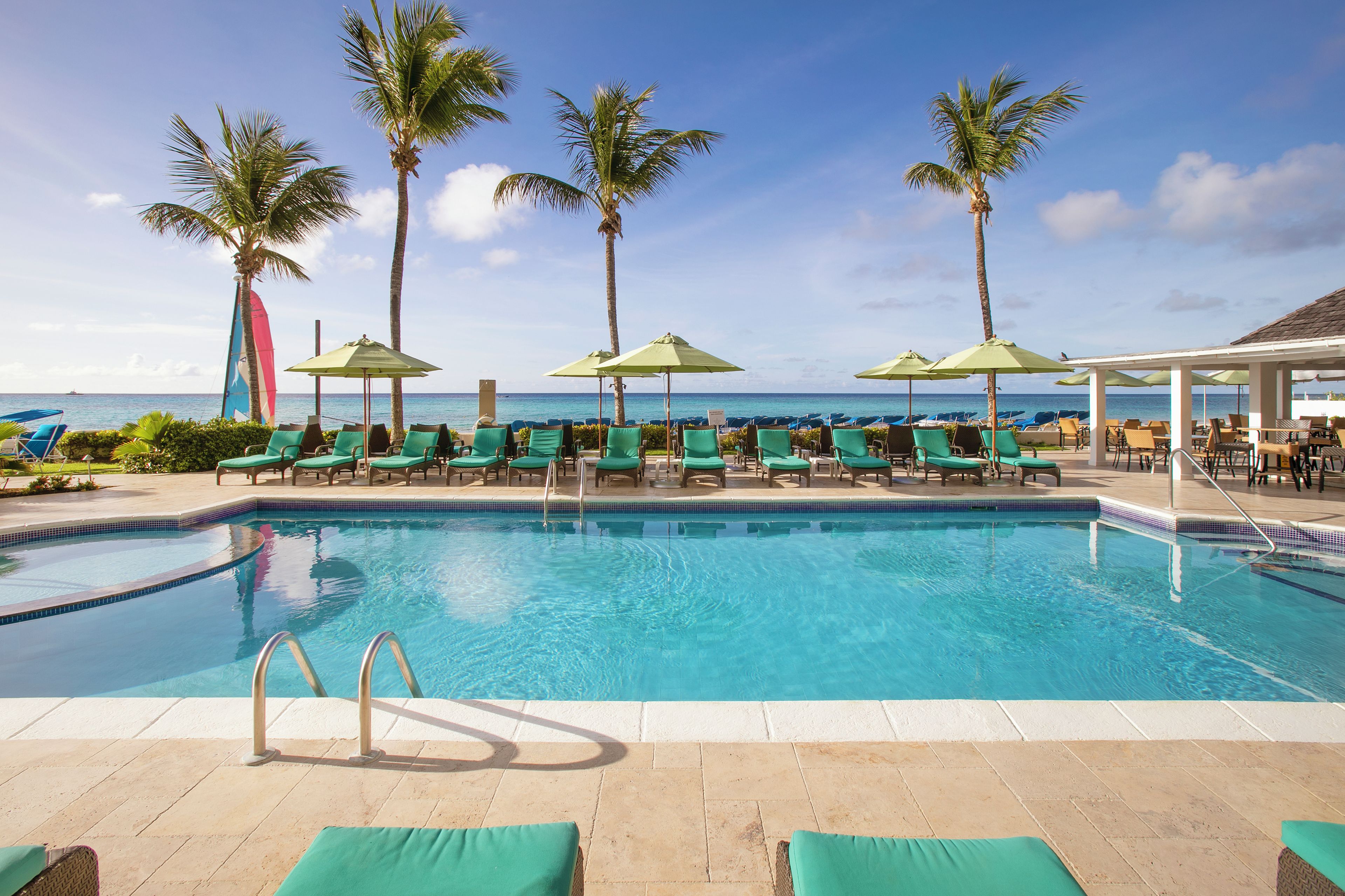 A poolside area with lounge chairs and umbrellas, palm trees, and a view of the ocean.