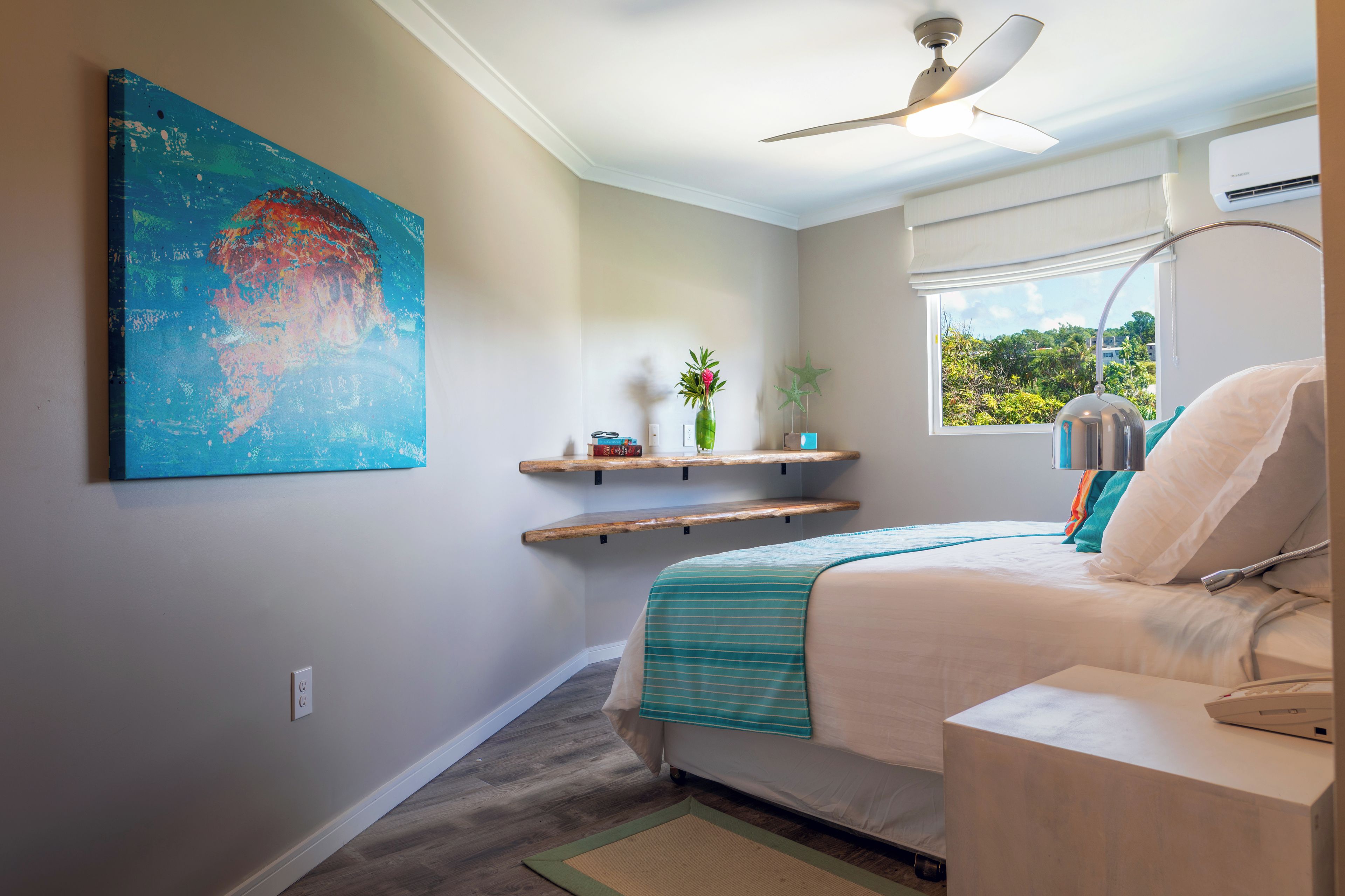 Un dormitorio con cama, ventilador de techo, ventana con vistas, un cuadro en la pared y una estantería con objetos.