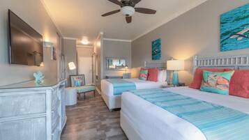 Een hotelkamer met twee bedden, een plafondventilator, kunstwerken aan de muur en een aan de muur bevestigde televisie.