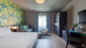 Ein modernes Hotelzimmer mit einem großen Bett, einer Couch, einem Sitzsack, einem Schreibtisch mit Stuhl, einem Fernseher und einem Fenster mit Vorhängen.