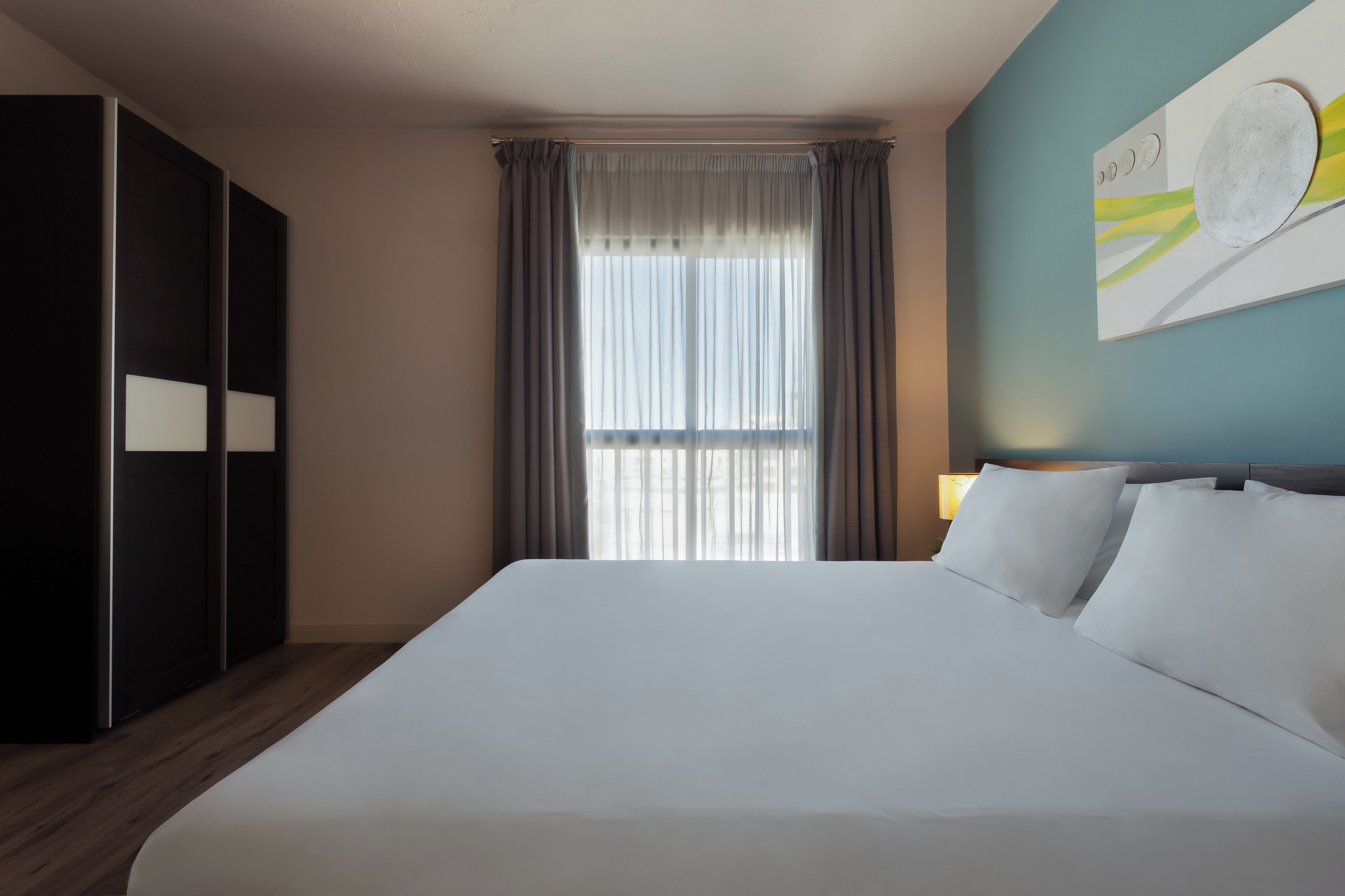 A hotel room with a large bed, a wardrobe, a window with curtains, and a wall with a painting.