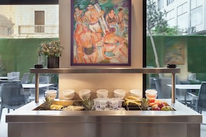 A buffet setup with bowls and fruits, a painting above, and outdoor seating visible through the window. - Argento Hotel Malta (St. Julian's)