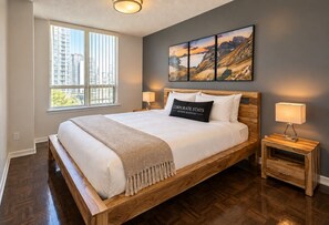 A bedroom with a large bed, two bedside tables, a window with a view of buildings, and a wall-mounted painting.