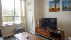 A living room with a large window, a flat-screen TV on a wooden stand, a patterned chair, and a small table with decorative items. - Stylish 1BR+Den Retreat in Markham (Markham)