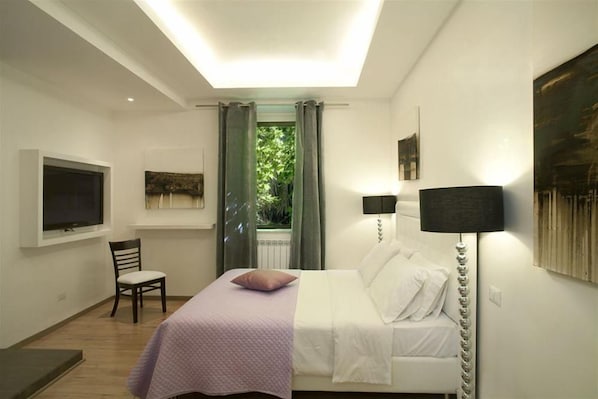 A bedroom with a bed, a chair, a TV, and a window with curtains.