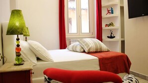 A bedroom with a bed, a nightstand, a lamp, a window with curtains, and a shelf with items.