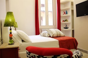 A bedroom with a bed, a nightstand, a lamp, a window with curtains, and a shelf with items.