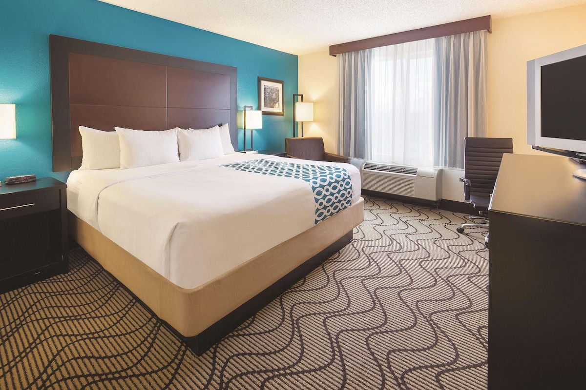 Quarto de hotel com uma cama grande, uma escrivaninha, uma cadeira, uma televisão e uma janela com cortinas.