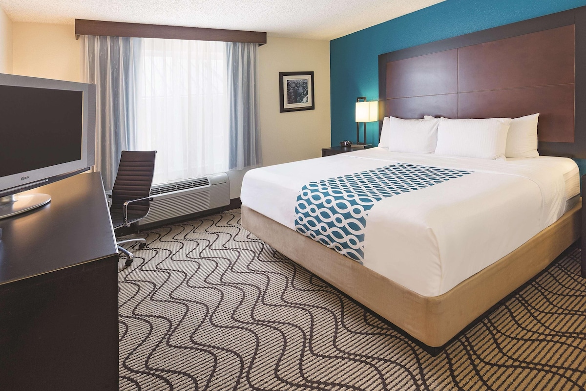 Quarto de hotel com uma cama, uma escrivaninha com televisão, uma cadeira, uma janela com cortinas, uma mesa de cabeceira com luminária e um tapete estampado.