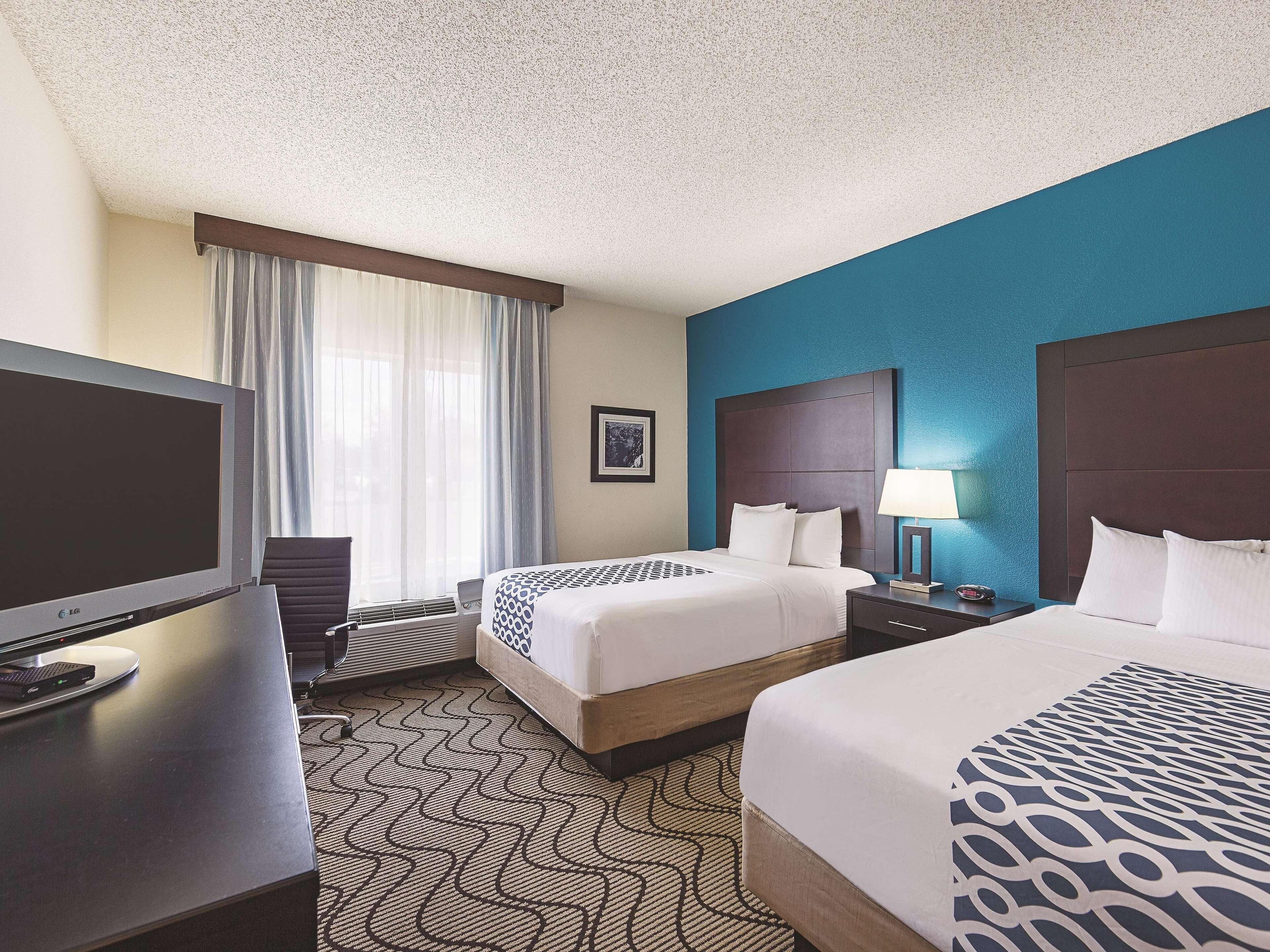 A hotel room with a large flat-screen TV, two beds with patterned bedspreads, a desk with a computer, and a window with curtains.