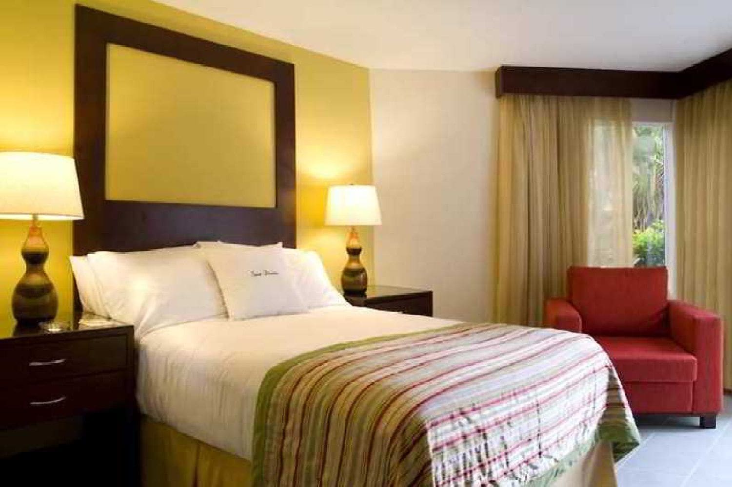 A hotel room with a bed, two bedside tables with lamps, a red armchair, and a large window with curtains.