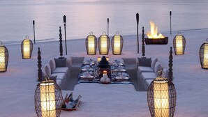 Vista del atardecer con una zona de comedor en la playa, que incluye una mesa larga con sillas, velas y un brasero.