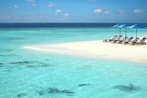 A beach with clear turquoise water, white sand, and blue umbrellas providing shade for lounge chairs.
