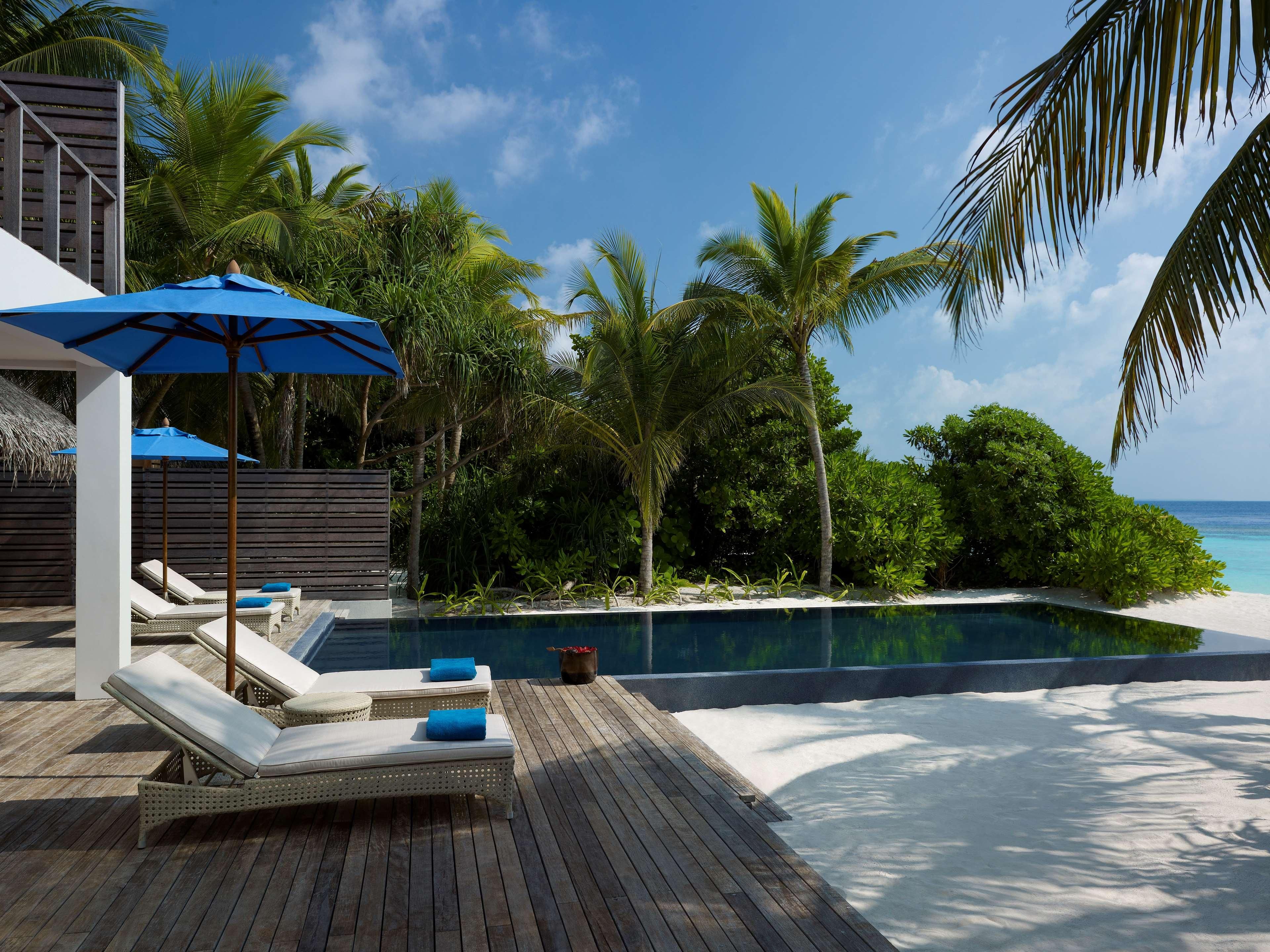 A poolside area with lounge chairs and a blue umbrella, surrounded by tropical vegetation and a clear view of the ocean.