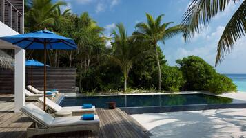 A poolside area with lounge chairs and a blue umbrella, surrounded by tropical vegetation and a clear view of the ocean.