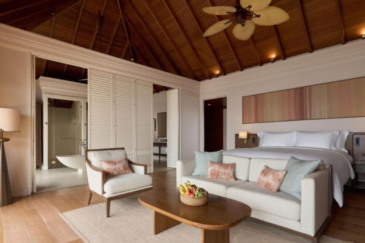 A spacious bedroom with a large bed, a sitting area with a chair and a table, and a ceiling fan.