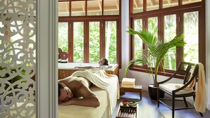 A spa room with a wooden ceiling, large windows, a massage table, a chair, a small table with a bowl of flowers, and a potted plant.