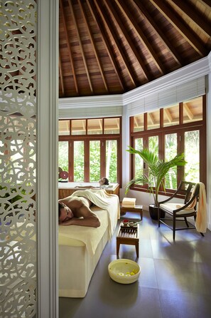 A spa room with a wooden ceiling, large windows, a massage table, a chair, a small table with a bowl of flowers, and a potted plant.